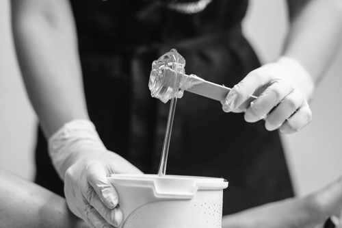 Frau mit einem kleinen Eimer Sugaring-Paste und etwas Paste an einem Holzstab. Im Hintergrund sieht man ein Frauenbein. Schwarz-Weiß-Bild